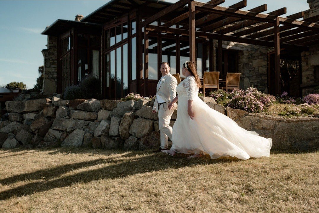 A bride and groom walking hand in hand across the lawn of a stone-and-timber Tasmanian property, the bride glancing back over her shoulder mid-stride
