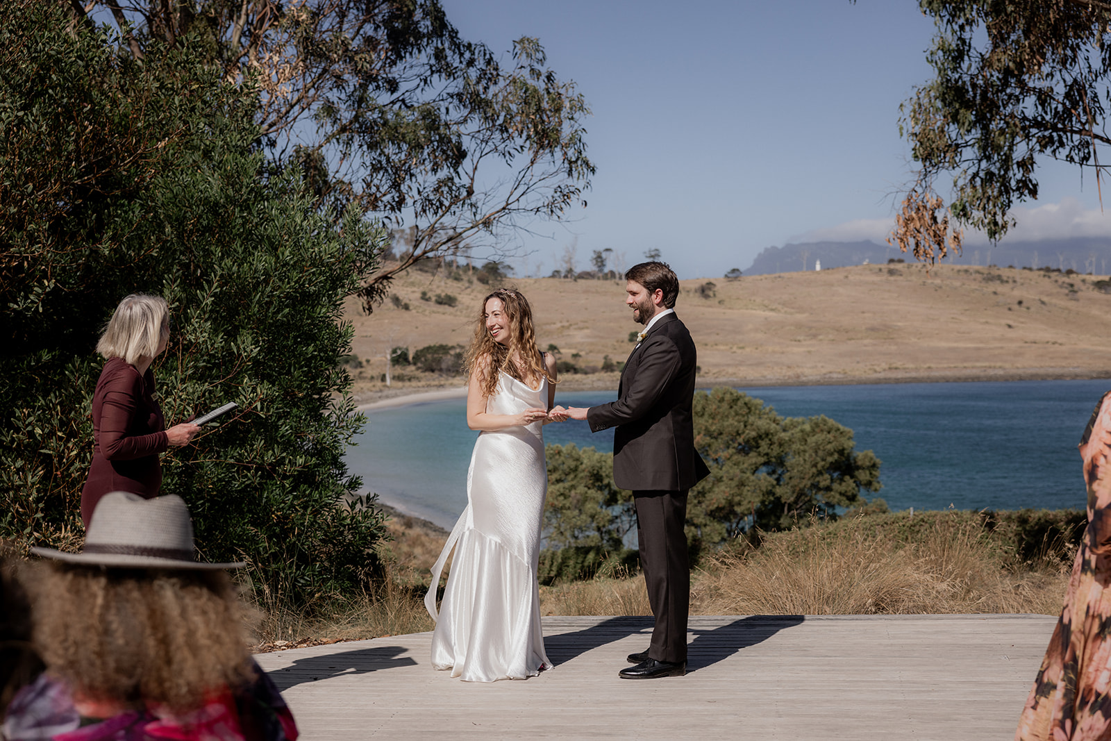 Mary Figg officiating a coastal wedding ceremony, with the bride and groom exchanging rings and guests seated nearby overlooking a Tasmanian bay