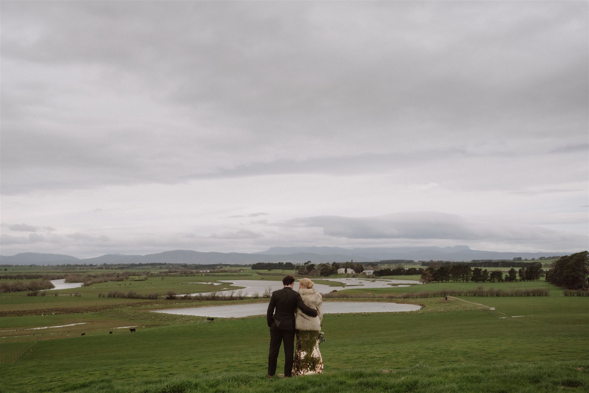 A newly married couple standing together on a grassy hillside overlooking farmland and distant Tasmanian mountains