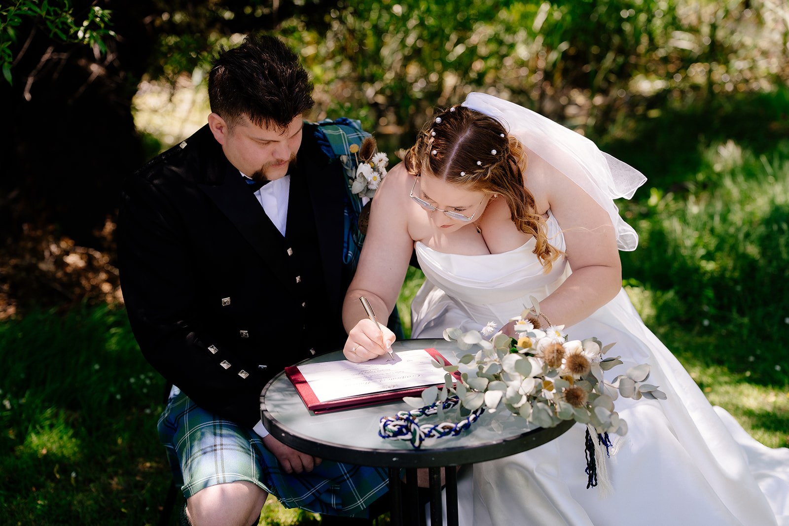 A bride leaning in to sign the marriage certificate at a small outdoor table, her groom seated beside her, with a bouquet and handfasting cord resting on the table and the wording out of focus